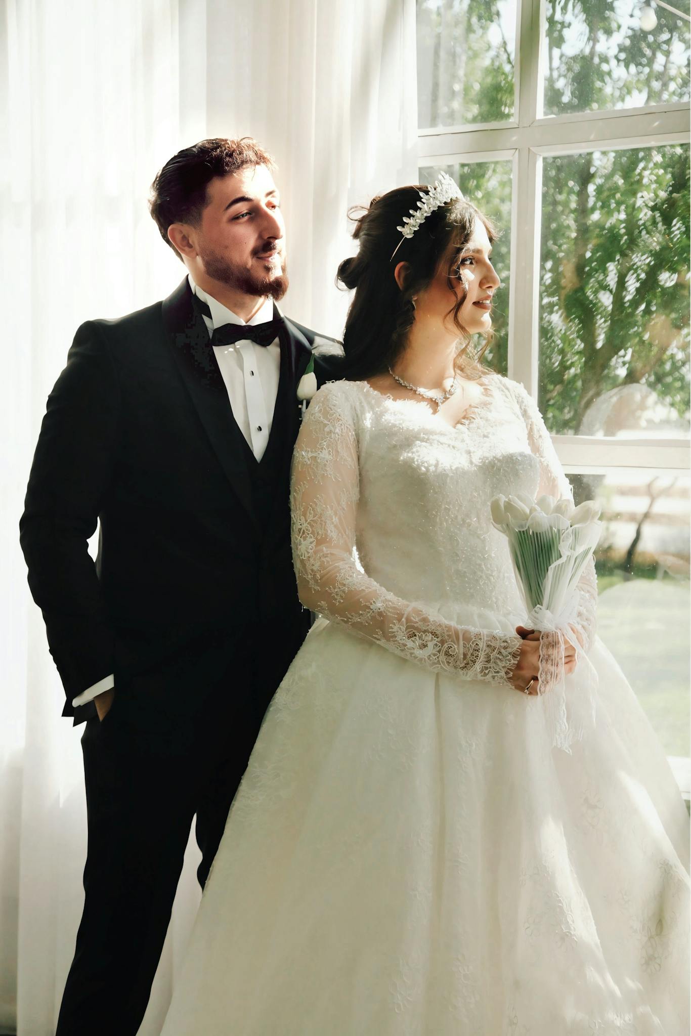 Bride and groom posing by a sunlit window, looking elegant and joyful.