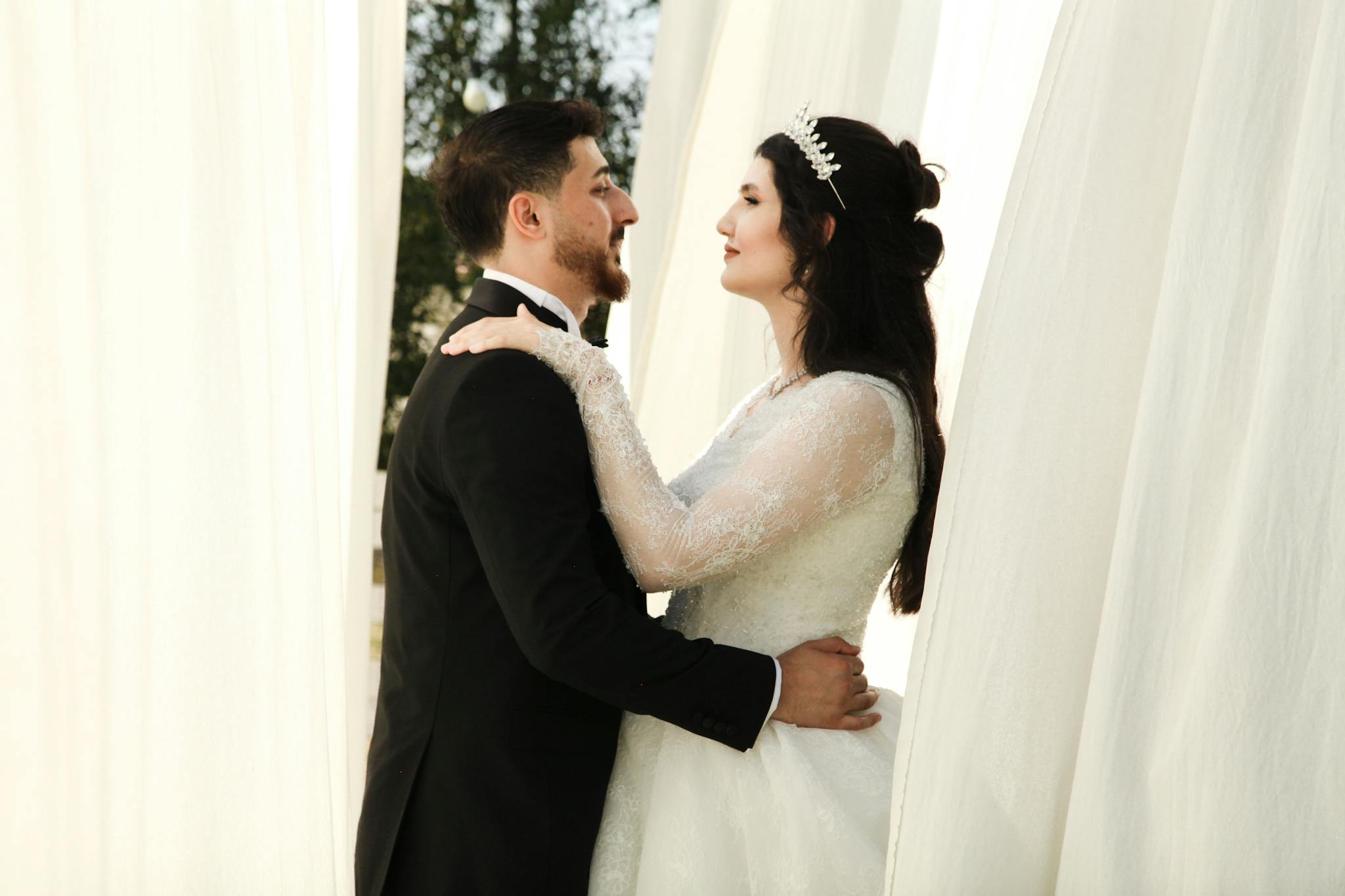 Elegant bride and groom embracing in wedding attire amidst flowing white curtains.